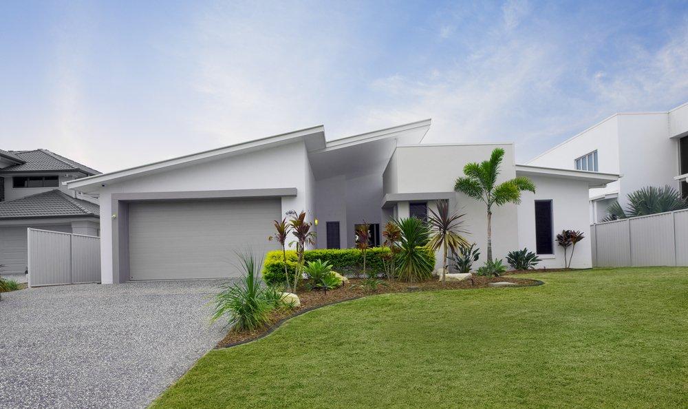 A modern white house with a slanted roof and a grey double garage door, fronted by a manicured lawn and tropical landscaping.