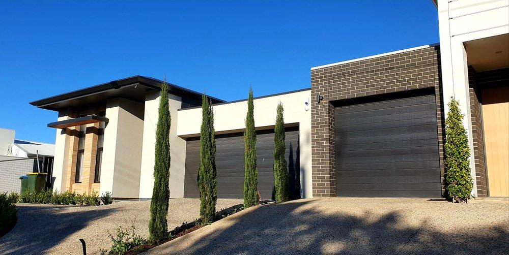 A modern residential house with light-coloured walls, dark brick accents, and black garage doors, featuring tall, slender cypress trees along the driveway.